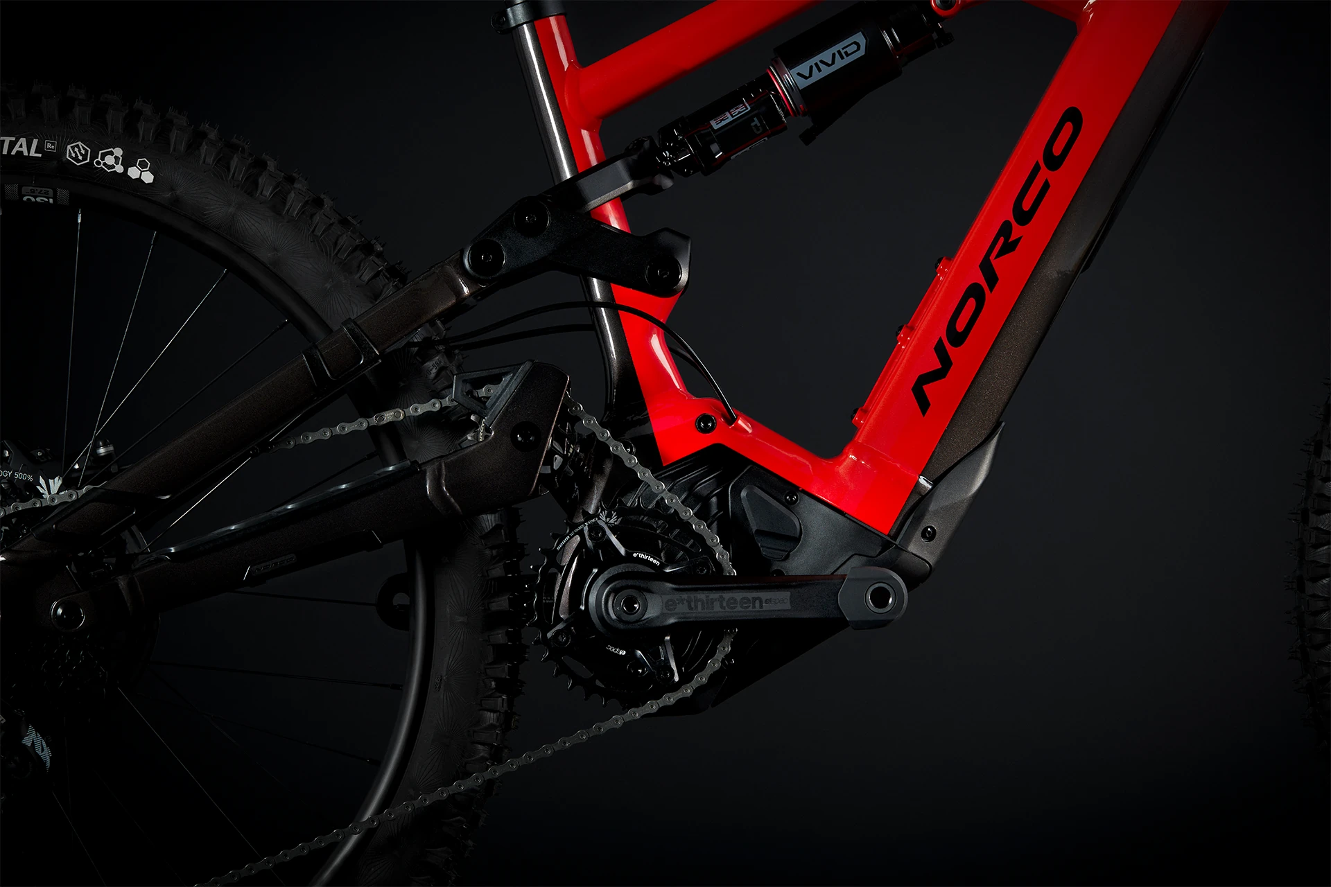 Gros plan d'un cadre de vélo de montagne Norco rouge, montrant la suspension arrière, la chaîne, la manivelle et une partie de la roue arrière sur un fond sombre.