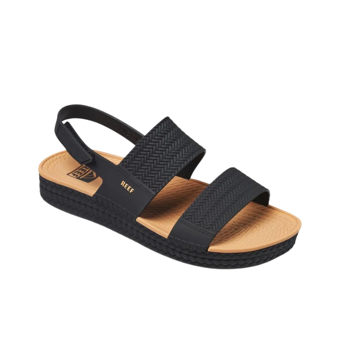 La WATER VISTA de REEF est une sandale noire et beige avec deux larges brides noires, une bride arrière, une semelle beige texturée, une plateforme noire à motifs et le logo "REEF" doré imprimé dessus.