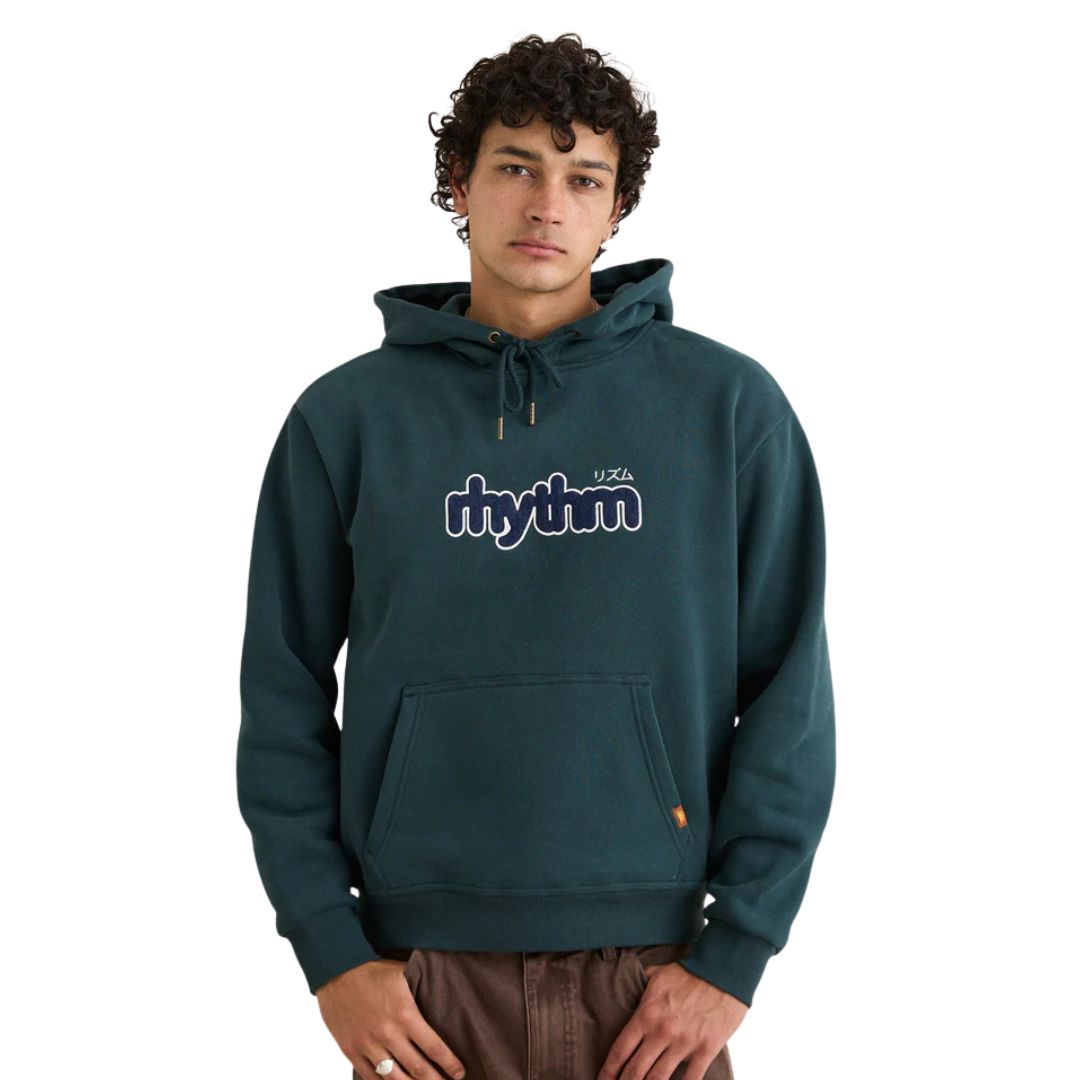 Un jeune homme aux cheveux bouclés porte le CHANDAIL À CAPUCHON EN MOLLETON RHYTHM APPLIQUE POUR HOMME, avec le mot "rhythm" en blanc et bleu marine sur le devant. Il se tient debout, sûr de lui, les mains dans les poches, en regardant la caméra.