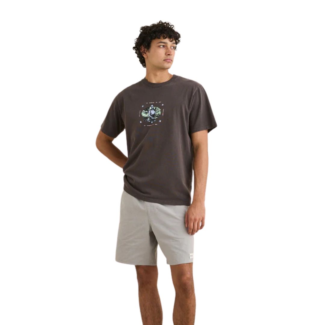 Un jeune homme aux cheveux bouclés porte le T-SHIRT BOTANICAL VINTAGE SS - un tee-shirt graphique foncé - associé à un short gris clair, debout, une main dans la poche et regardant sur le côté, sur un fond blanc uni.