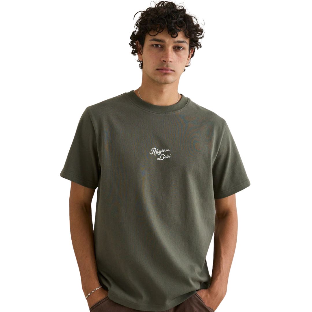 Un jeune homme aux cheveux bruns bouclés porte le T-SHIRT RHYTHM HEAVY VINTAGE BRODÉ POUR HOMME, un tee-shirt vert olive avec "Requiem Paris" en petites lettres blanches sur la poitrine, debout, les mains dans les poches sur un fond blanc uni.