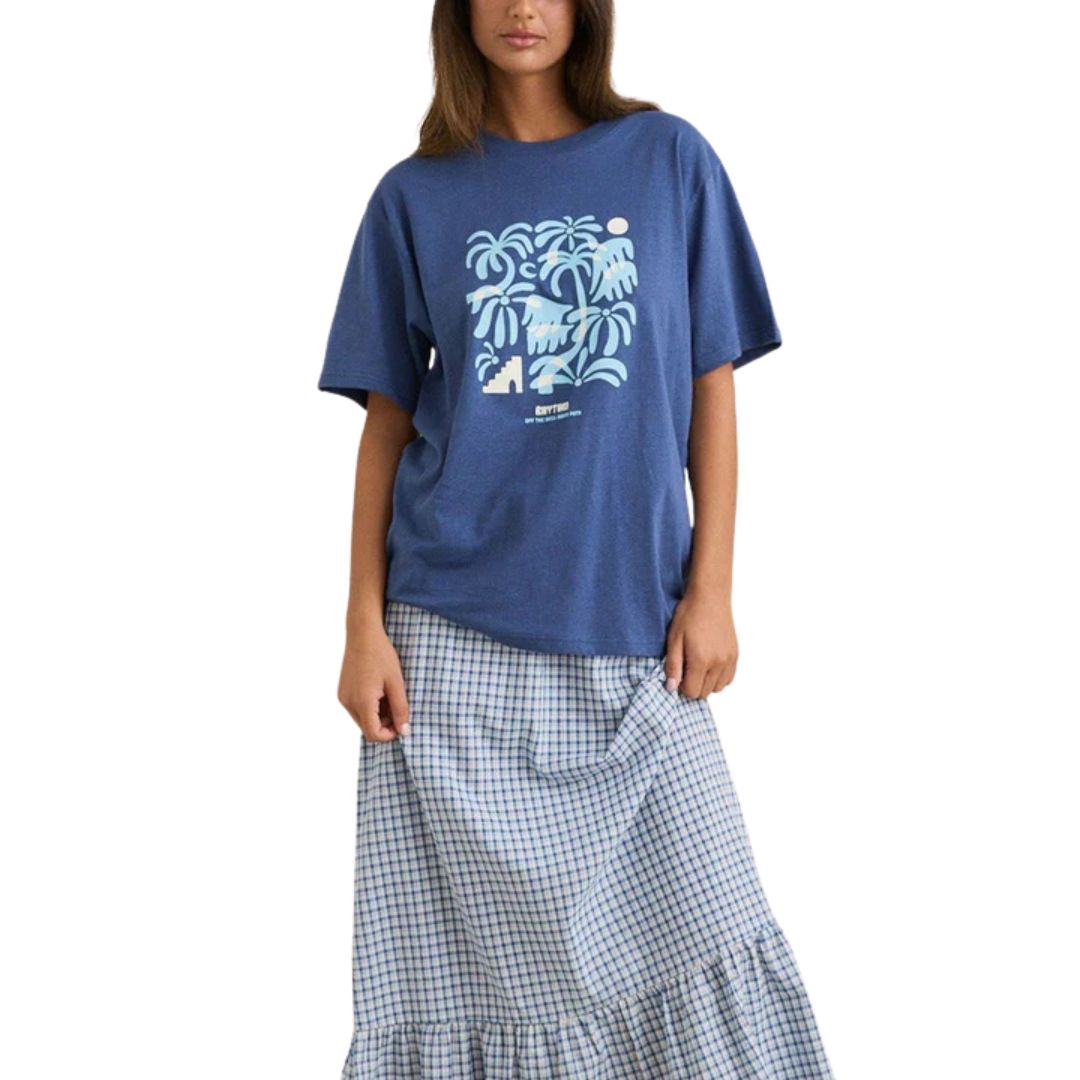 Une femme porte le ISLAND OVERSIZED TEE, un T-shirt graphique bleu avec un motif tropical blanc, associé à une longue jupe à carreaux bleus et blancs, sur fond blanc.