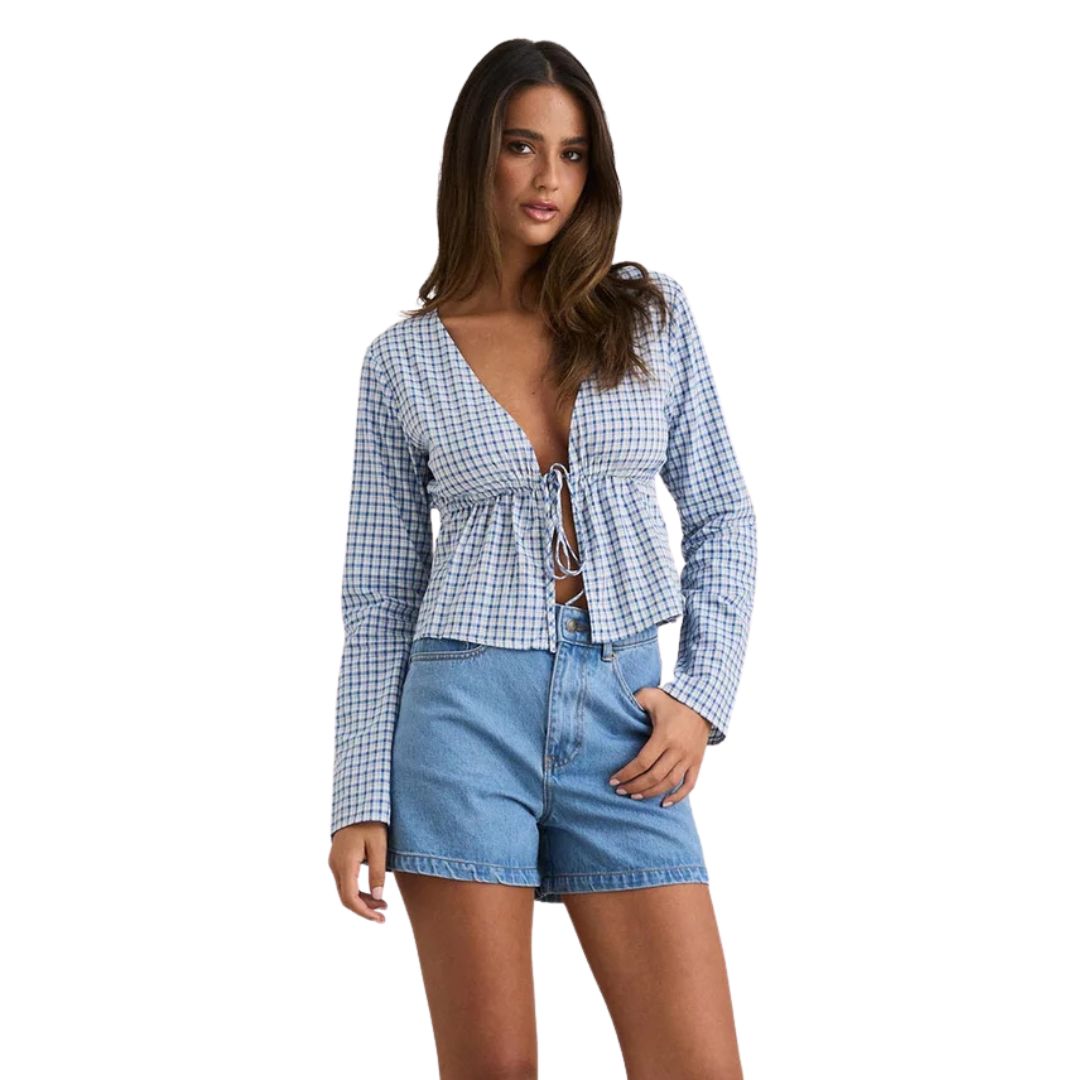 Une femme aux cheveux longs et lâchés pose dans le HAUT À NOUER À MANCHES LONGUES RHYTHME LOLA POUR FEMME, un crop top à manches longues à carreaux bleus et blancs avec cravate sur le devant, associé à un short en jean bleu à taille haute sur fond blanc.