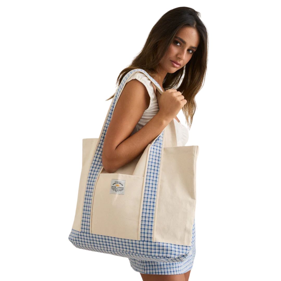 Une femme pose sur un fond blanc uni, tenant le LOLA CHECK TOTE BAG - un grand sac fourre-tout beige avec des accents écossais bleus et une petite étiquette - tout en portant un haut blanc sans manches et un short rayé.