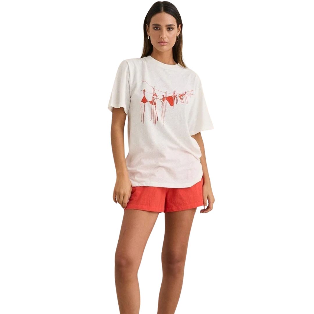 Une femme porte le T-SHIRT SURDIMENSIONNÉ RHYTHM MELIA POUR FEMME - un tee-shirt blanc surdimensionné avec un graphisme rouge - associé à un short rouge, sur fond blanc et incarnant un style décontracté et sans effort.