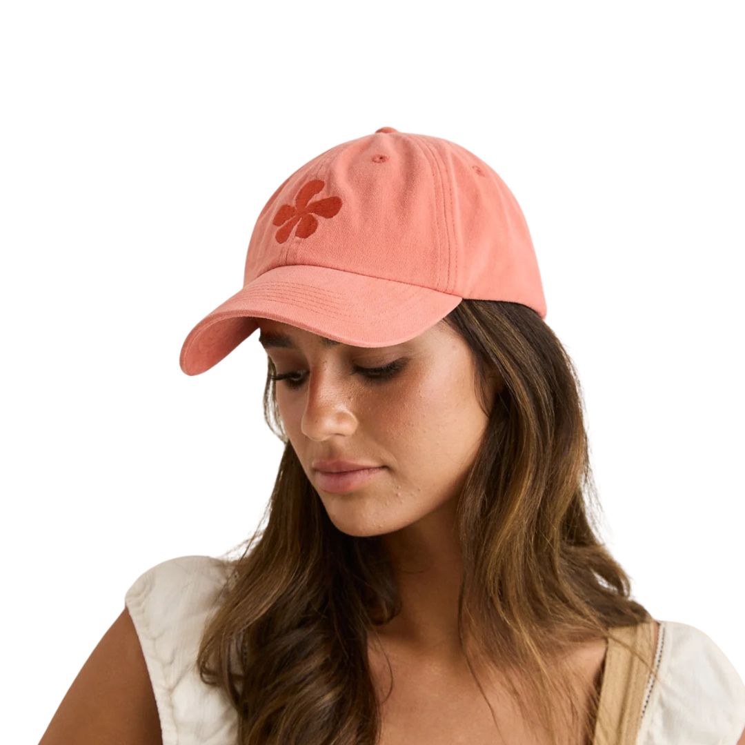 Une femme aux longs cheveux bruns porte la casquette MELIA FLORAL CAP, une casquette de baseball rose corail ornée d'un trèfle à quatre feuilles rouge. Elle regarde vers le bas en souriant légèrement et est vêtue d'un haut blanc avec une bretelle beige sur l'épaule.