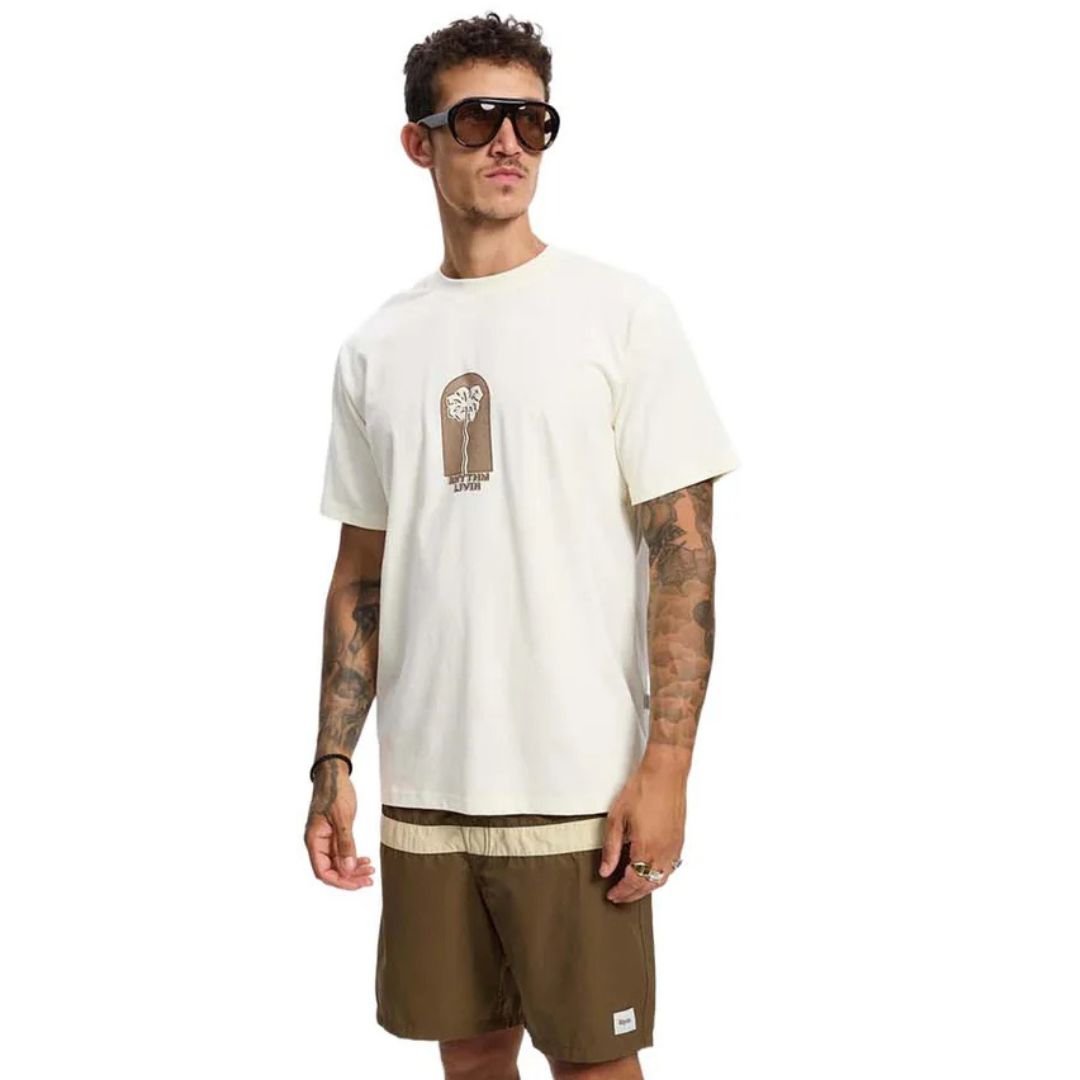 Un homme tatoué, portant de grandes lunettes de soleil, vêtu du T-SHIRT SHAKEN VINTAGE SS (crème avec un motif graphique) et d'un short marron, se tient debout sur un fond blanc uni.
