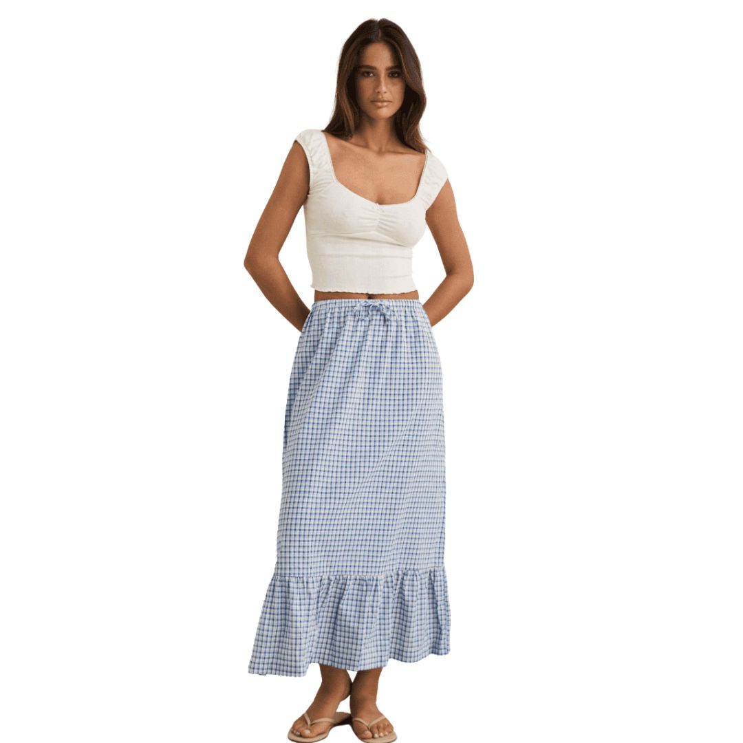 Une femme aux longs cheveux bruns, les mains derrière le dos, porte la JUPE LONGUE À VOLANTS RHYTHM MAXI LOLA POUR FEMME - une jupe maxi à volants vichy bleu et blanc - avec un haut à manches capuchon ajusté blanc et des sandales sur fond blanc.