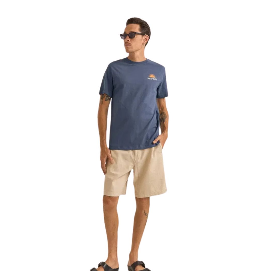 Un homme portant des lunettes de soleil, le TEE AWAKE (un T-shirt bleu avec un petit graphique sur la poitrine), un short beige et des sandales noires se tient sur un fond blanc.