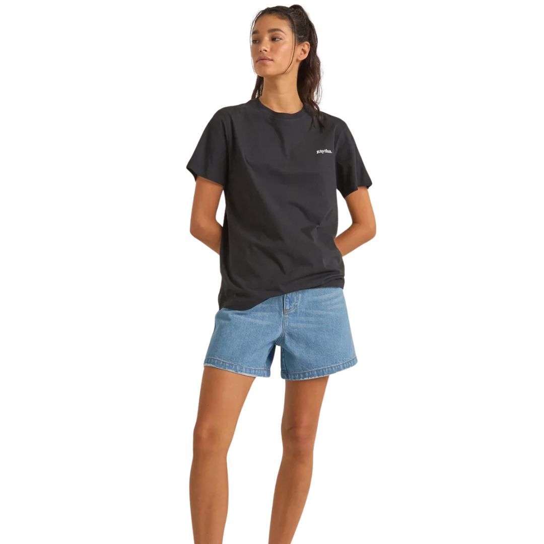 Une femme aux longs cheveux bruns tirés en arrière se tient debout, les mains derrière le dos, vêtue du T-SHIRT LOGO BOYFRIEND en noir et d'un short en jean bleu. Elle regarde légèrement sur le côté sur un fond blanc uni.