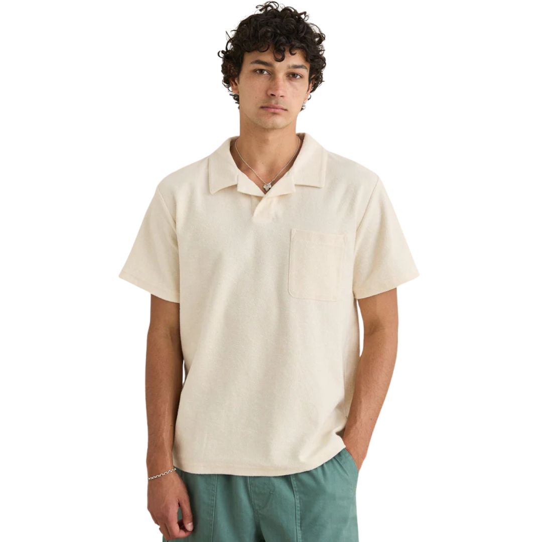 Un jeune homme aux cheveux bouclés se tient debout, les mains dans les poches, regardant la caméra. Il porte un pantalon vert et le VINTAGE TERRY POLO de couleur crème avec des manches courtes et une poche sur la poitrine.