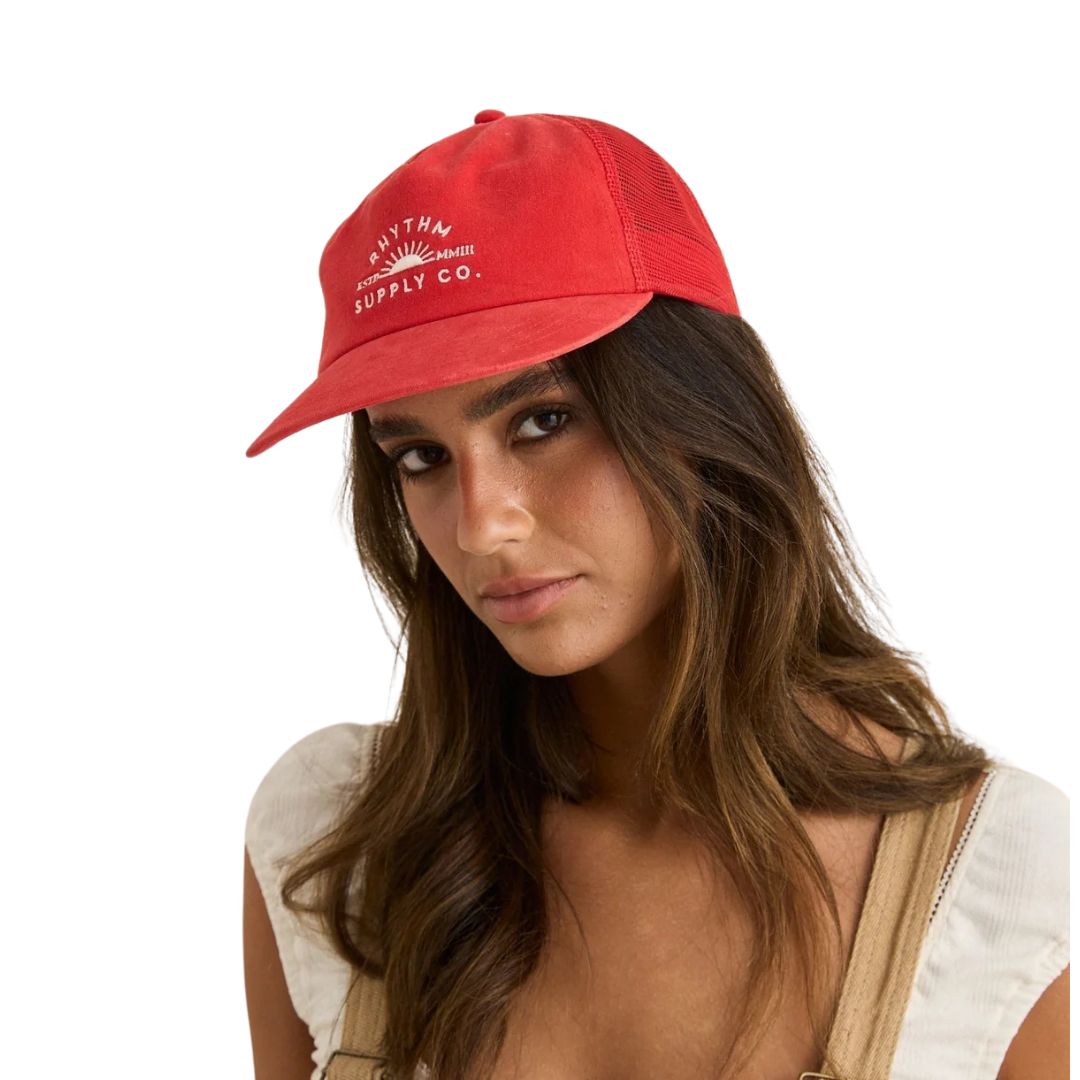 Une femme aux longs cheveux bruns porte la casquette RHYTHM SUPPLY TRUCKER CAP, avec un motif rouge et une marque sur le devant. Elle l'associe à un haut blanc et à une salopette beige, regardant l'appareil photo de manière neutre sur un fond blanc.