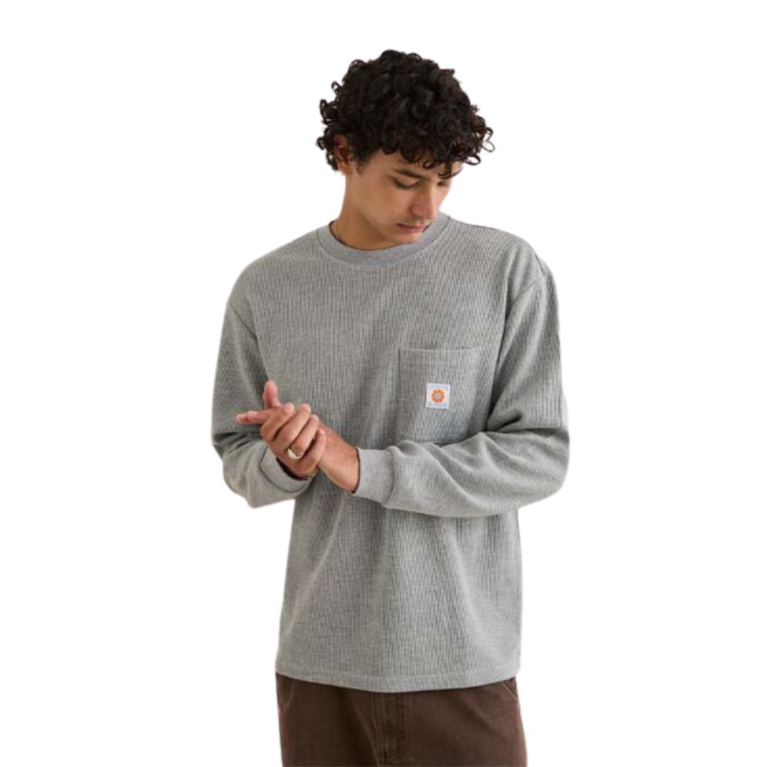 Une personne aux cheveux bouclés porte le VINTAGE POCKET LS WAFFLE, une chemise grise à manches longues avec une poche avant et un petit logo blanc, debout sur un fond blanc et regardant vers le bas tout en touchant ses mains.