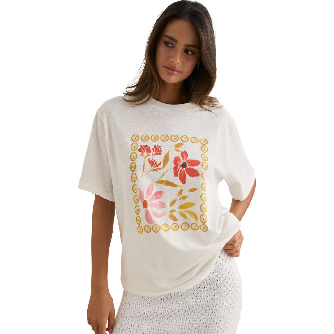 Une femme porte le T-SHIRT OVERSIZED WILDFLOWER, avec un imprimé floral coloré, associé à une jupe blanche texturée. Elle se tient debout sur un fond blanc uni, regardant sur le côté et tenant son tee-shirt d'une main.