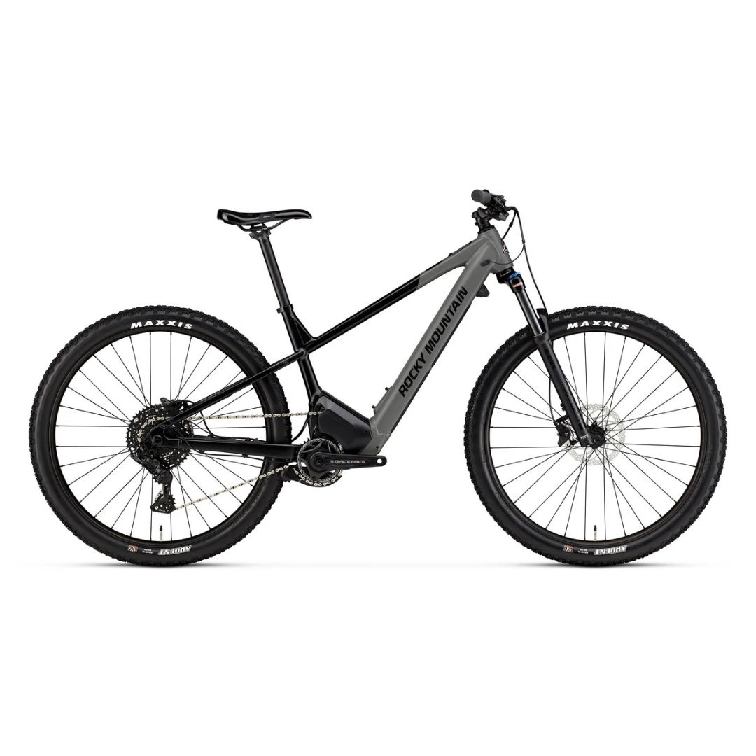 Un vélo de montagne électrique Rocky Mountain noir et gris avec un cadre élégant, une fourche à suspension avant, des freins à disque et de gros pneus Maxxis à crampons, présenté en vue latérale sur fond blanc.