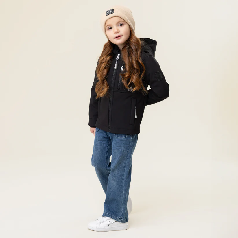 Un jeune enfant se tient debout, confiant, vêtu d'une veste noire, d'un jean bleu, de baskets blanches et d'un bonnet beige. L'arrière-plan, d'une couleur simple et claire, met en valeur la pose et la tenue de l'enfant.