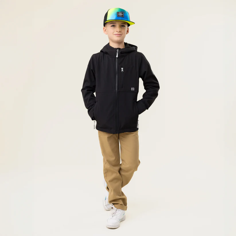 Un jeune homme vêtu d'un sweat à capuche noir, d'un pantalon beige et d'une casquette colorée se tient debout, les mains dans les poches, l'air confiant. Le fond est beige clair uni.