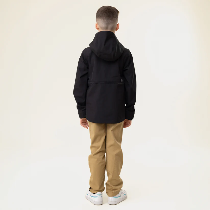 Un jeune garçon se tient dos à l'objectif, vêtu d'un sweat à capuche noir, d'un pantalon kaki et de baskets blanches aux accents colorés. L'arrière-plan est uni et blanc cassé.