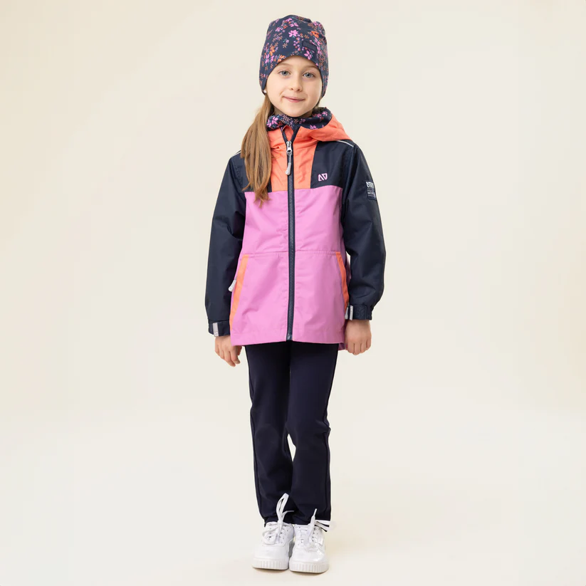 Une jeune fille se tient debout sur un fond neutre, vêtue d'une tenue d'hiver colorée composée d'une veste rose et orange, d'un pantalon bleu et d'un chapeau à motifs floraux. Elle porte des chaussures blanches et ses cheveux sont détachés, laissant dépasser des mèches de son chapeau.