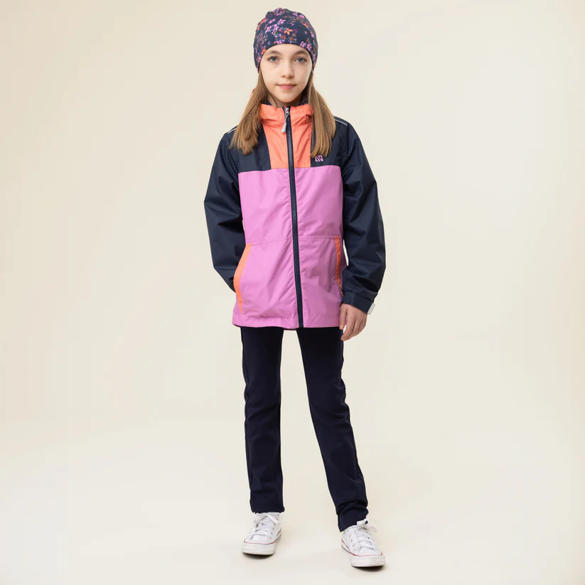 Une jeune fille portant une veste colorée avec des panneaux bleu marine, rose et orange, un pantalon foncé, des baskets blanches et un bonnet à motifs floraux se tient sur un fond uni.