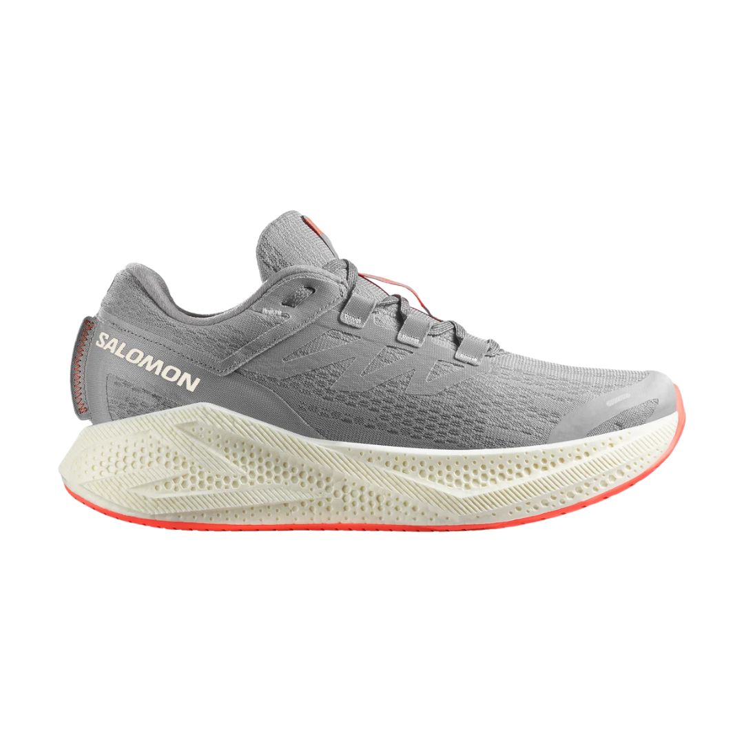 Une chaussure de course Salomon grise avec des accents blancs et orange, une tige en mesh texturé et une semelle épaisse en mousse blanc cassé avec une bande de roulement orange subtile.