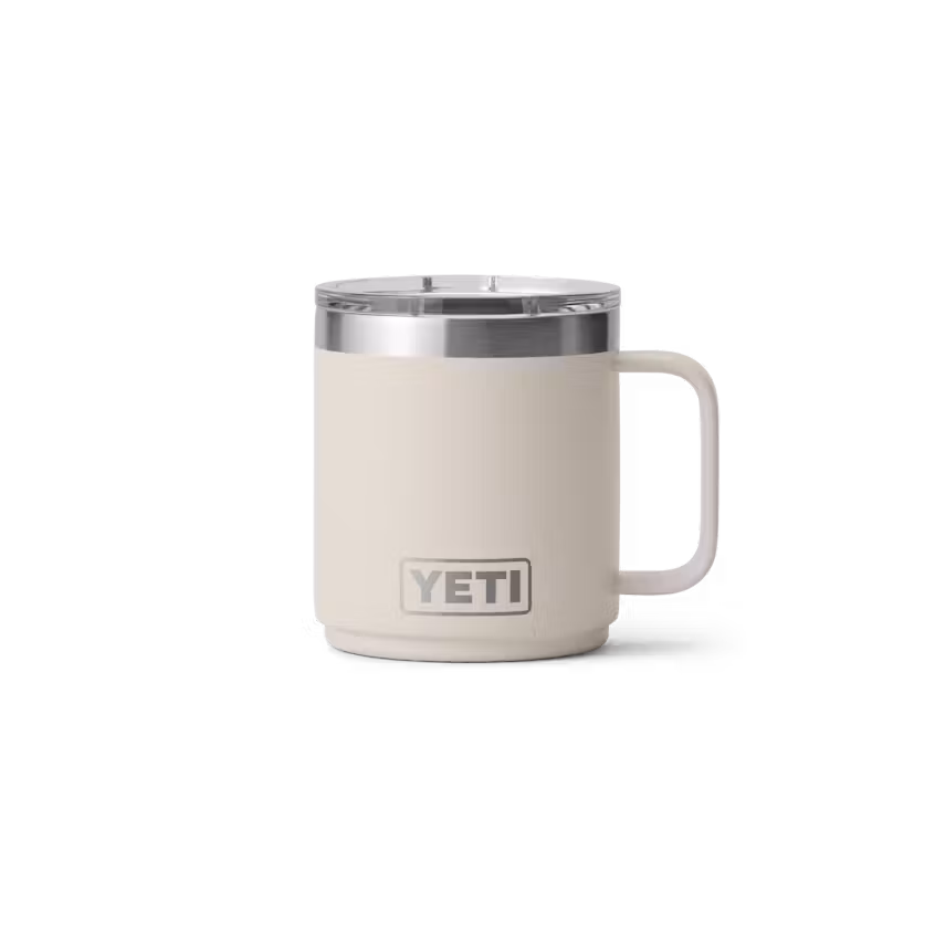 Une tasse RAMBLER 10 OZ MUG MS de YETI, de couleur beige, dotée d'une anse et d'un couvercle en acier inoxydable, est présentée sur un fond blanc uni. Le logo YETI figure sur le devant de la tasse.
