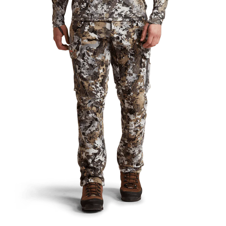 Une personne porte un pantalon de chasse à motif camouflage associé à des bottes de randonnée. La personne est représentée des épaules aux pieds, les mains posées le long des jambes. L'arrière-plan est noir, ce qui met en valeur la tenue.