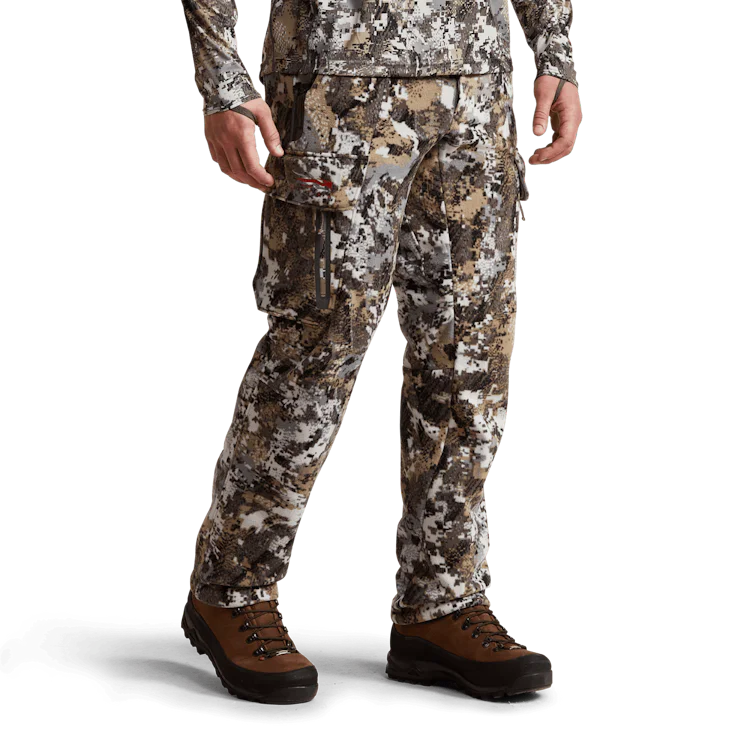 Une personne porte un pantalon de camouflage et un haut à manches longues assorti sur fond uni. Le pantalon a des poches cargo et la personne porte également des bottes de randonnée marron.