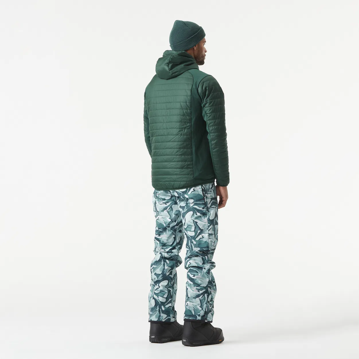 Une personne se tient debout, le dos tourné, vêtue d'une veste vert foncé, d'un bonnet et d'un pantalon à motifs bleus avec des bottes de neige. L'arrière-plan est blanc uni.