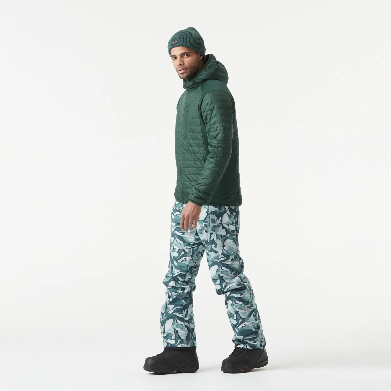 Un homme portant une veste à capuche verte, un pantalon camouflage et des bottes noires se tient debout sur un fond uni. Il regarde de côté, légèrement tourné vers la caméra. Il porte également un bonnet.