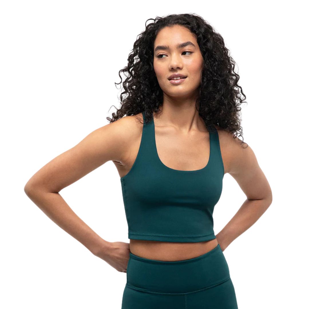 Une femme aux cheveux bouclés portant un haut court sans manches vert foncé et un legging taille haute assorti se tient debout, les mains sur les hanches, regardant sur le côté sur un fond blanc uni.