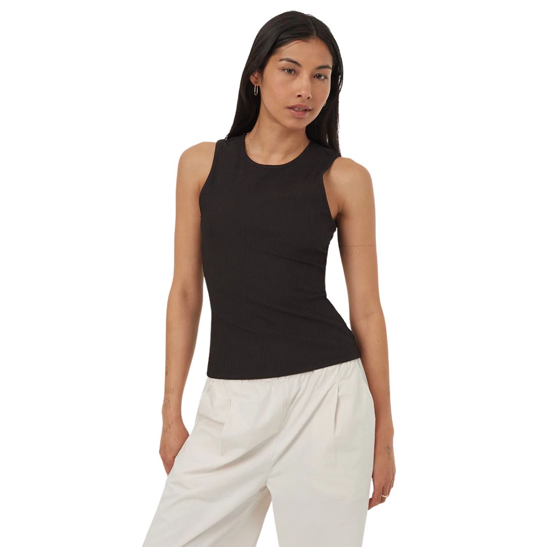 Femme aux longs cheveux noirs portant un haut noir sans manches et un pantalon blanc ample, debout sur un fond blanc uni.