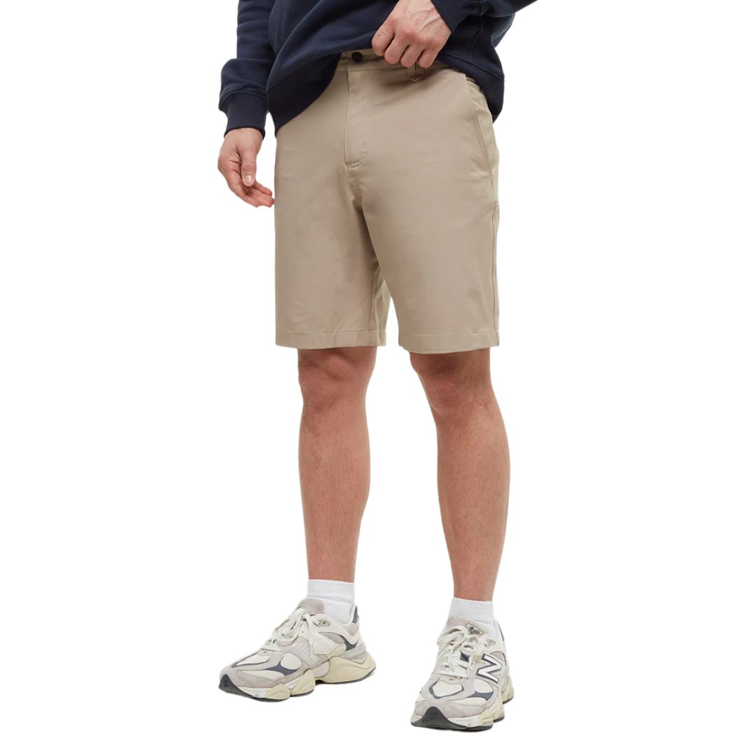 Une personne portant un sweat-shirt bleu marine, un short beige jusqu'aux genoux, des chaussettes blanches et des baskets beiges se tient debout avec une main dans sa poche sur un fond uni.