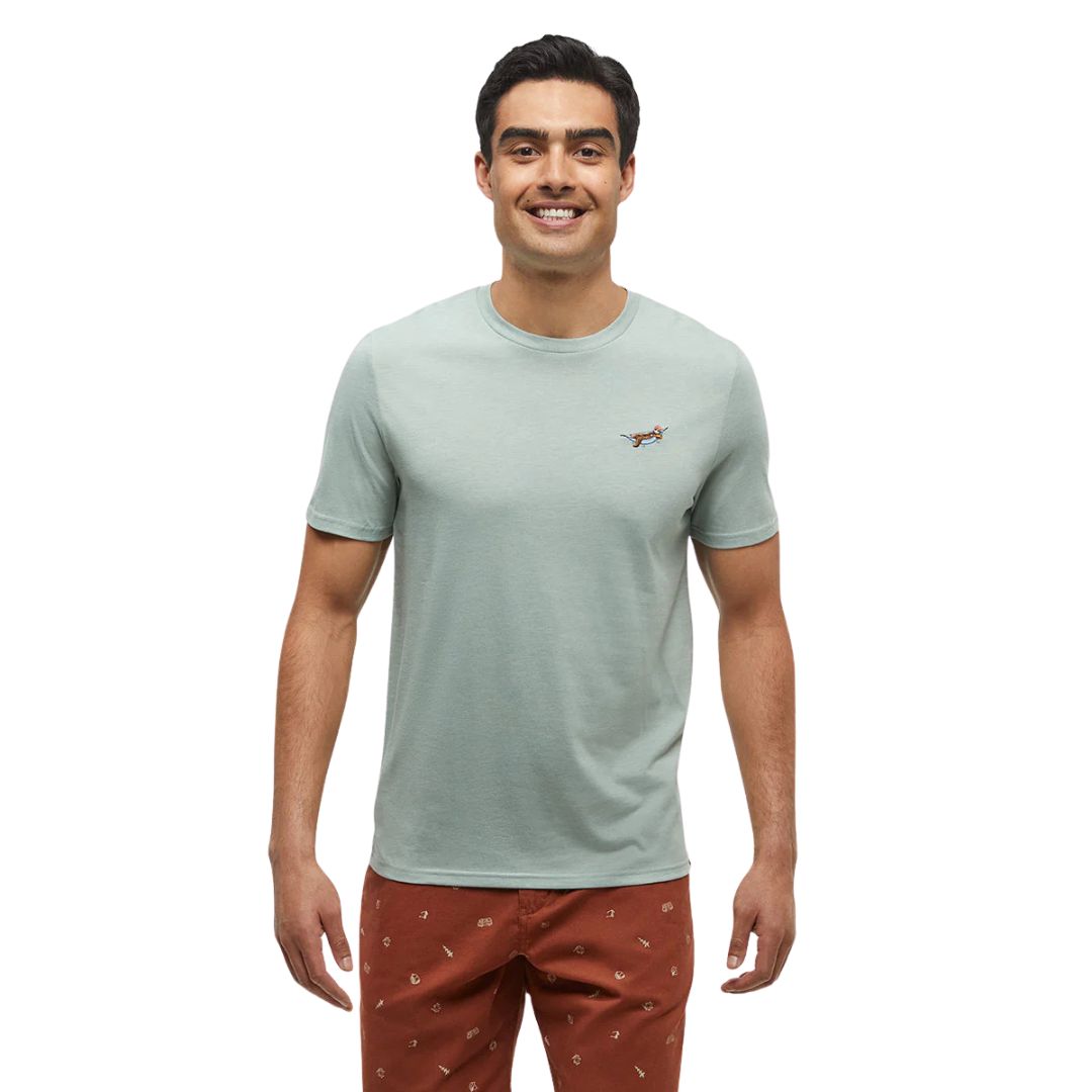 Un homme aux cheveux noirs courts sourit et se tient debout sur un fond uni. Il porte un t-shirt vert clair orné d'un petit motif brodé sur la poitrine et un pantalon brun-rouge à motif discret.