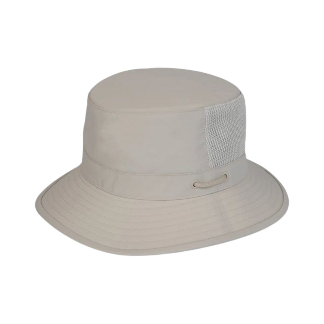 Le CHAPEAU BUCKET TILLEY AIRFLO BUCKET LTM1 est un chapeau baquet beige clair avec un large bord, un panneau d'aération latéral en maille et un œillet métallique pour la circulation de l'air, sur fond blanc.