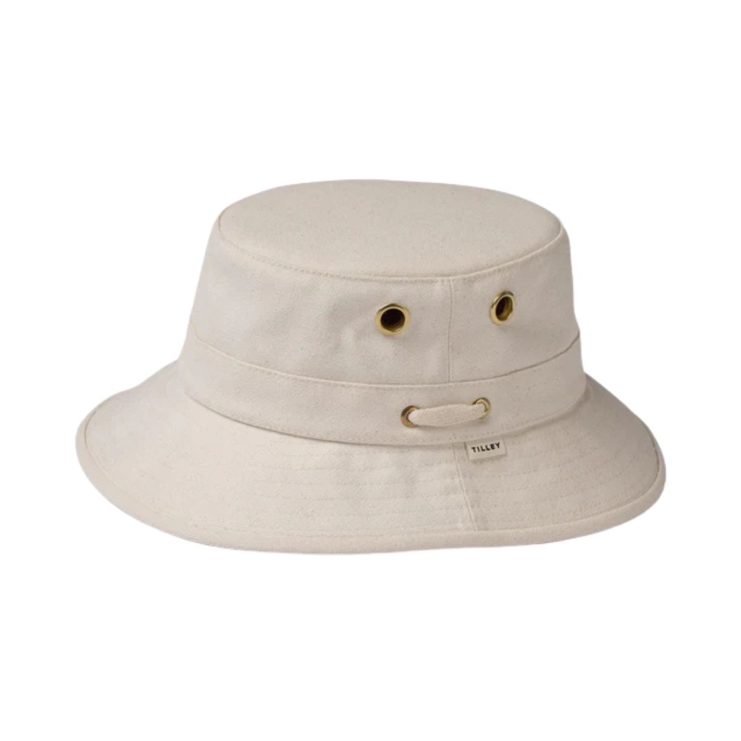 Le CHAPEAU TILLEY ICONIQUE T1 est un chapeau baquet beige avec un large bord, deux œillets métalliques de chaque côté et une petite étiquette en tissu sur la bande. Fabriqué en tissu robuste, il présente un design simple et classique.