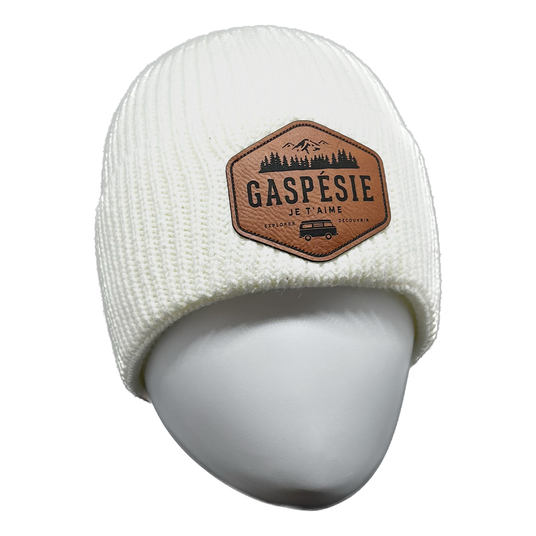 Tuque en tricot blanc avec un patch hexagonal brun sur le devant, comportant les mots « GASPÉSIE » et « Je t'aime » accompagnés d'une illustration de montagnes, d'arbres et d'un véhicule. Le produit s'appelle TUQUE EN TRICOT GASPÉSIE JE T'AIME LOGO VAN UNISEXE ADULTE.