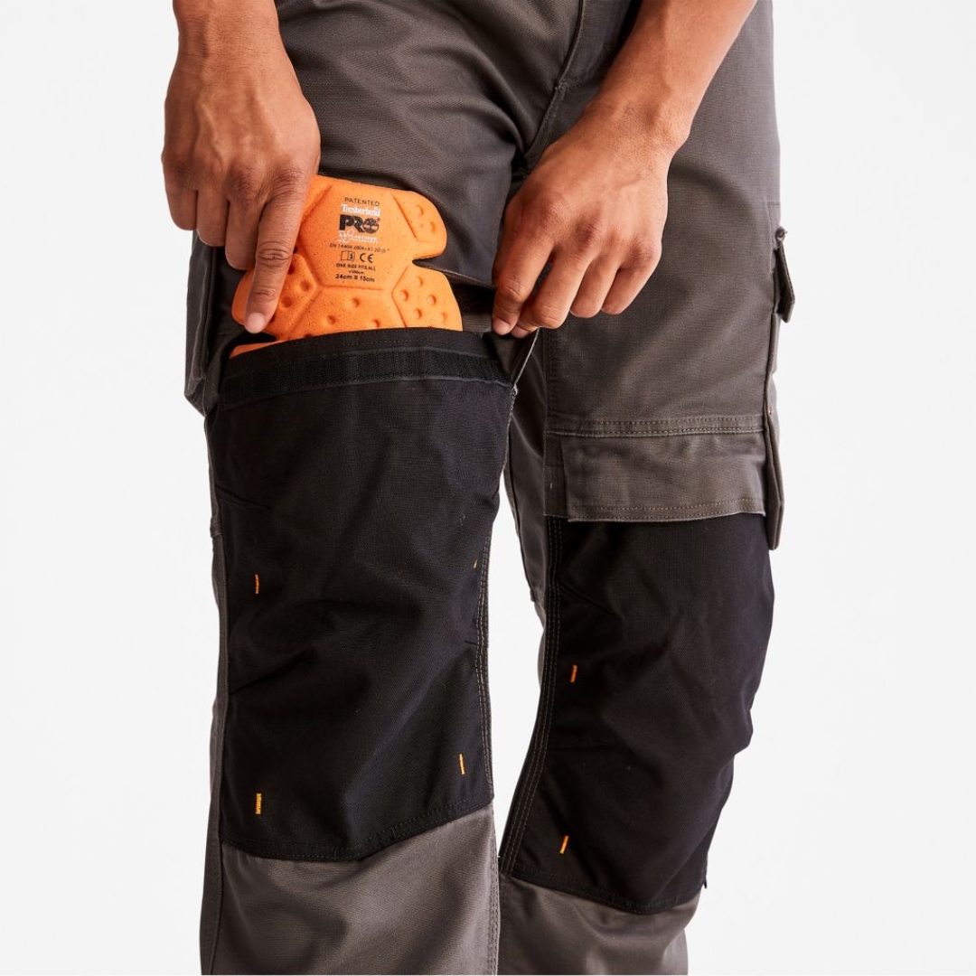 PANTALON DE TRAVAIL TIMBERLAND PRO IRONHIDE FLEX À GENOULLIÈRES POUR HOMME couleur gunmetal porté par un homme qui insert une genoullière anti fatigue dans la poche prévue à cette fin aux genoux du pantalon gris et noir