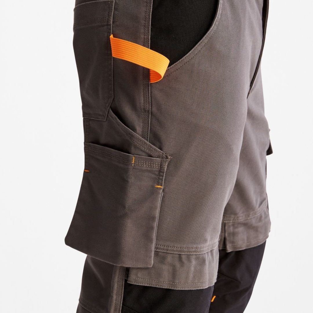 PANTALON DE TRAVAIL TIMBERLAND PRO IRONHIDE FLEX À GENOULLIÈRES POUR HOMME couleur gunmetal porté par un homme vu de coté gros plan sur la cuisse droite, ganse orangée pour le marteau visible, poche à vis, poche latérale pour les mains ainsi que la poche arrière visibles sur le pantalon gris acier