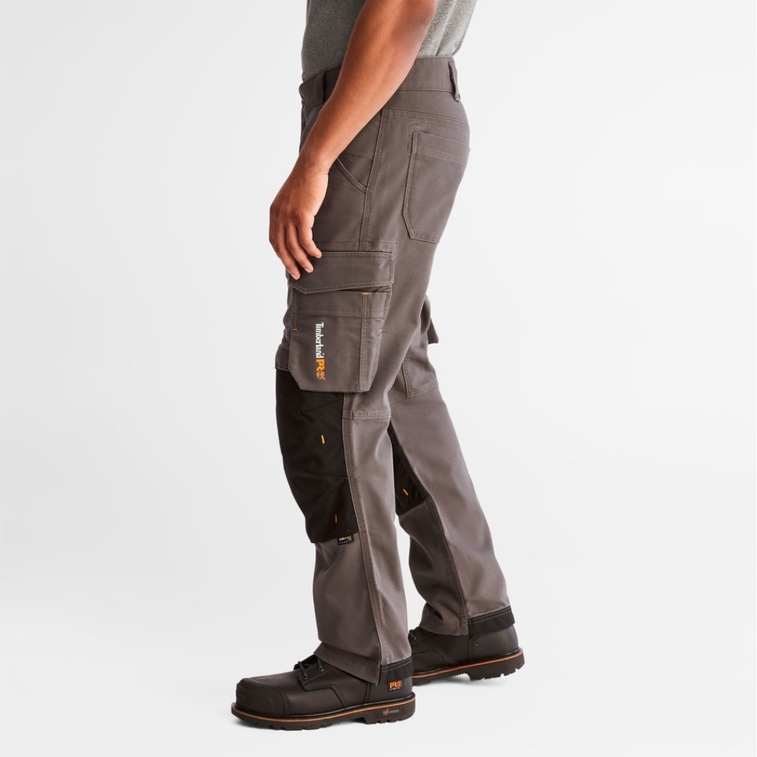 PANTALON DE TRAVAIL TIMBERLAND PRO IRONHIDE FLEX À GENOULLIÈRES POUR HOMME couleur gunmetal porté par un homme vu de profil gauche logo timberland pro imprimé sur la poche cargo visible, renforts noirs à l'ourlet et aux genoux visibles