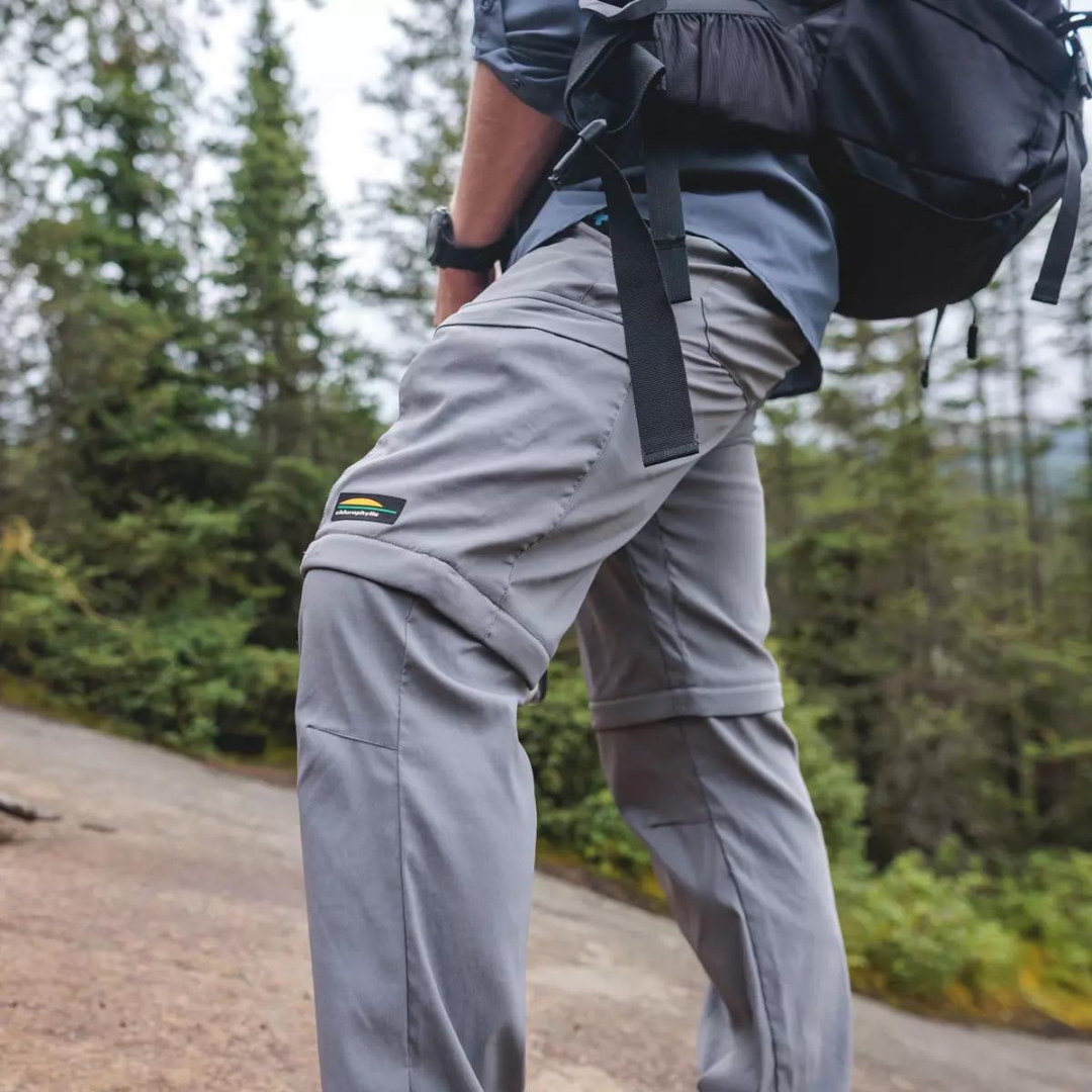 PANTALON DE PLEIN AIR CONVERTIBLE CHLOROPHYLLE RAID POUR HOMME COULEUR GRIS PIERRE PORTÉ PAR UN HOMME EN RANDONNÉE LOGO CHLOROPHYLLE SUR LA CUISSE GAUCHE VISIBLE,