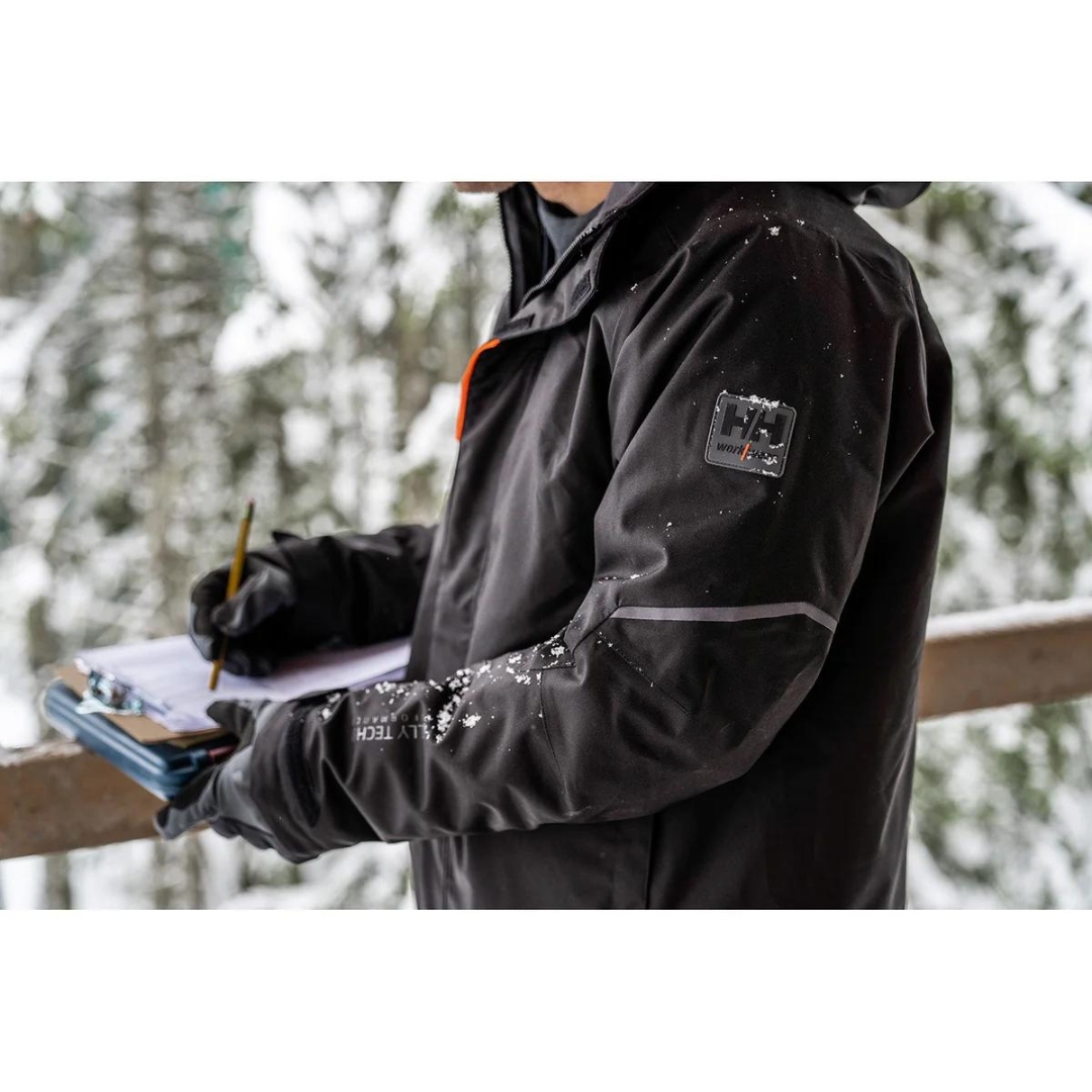 MANTEAU DE TRAVAIL ISOLÉ HELLY HANSEN WORKWEAR KENSINGTON POUR HOMME couleur black vu du manteau noir avec détails orange fluo porté par un homme vu dehors en gros plan sur la manche du manteau