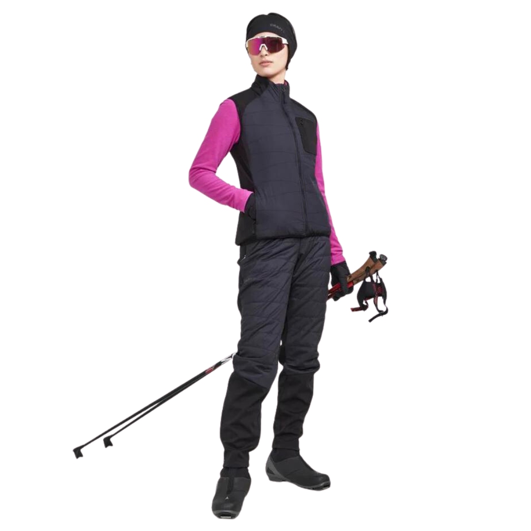 PANTALON ISOLÉ DE SKI DE FOND CRAFT CORE NORDIC TRAINING INSULATE POUR FEMME COULEUR BLACK VU DU PANTALON NOIR PORTÉ PAR UNE FEMME VUE DE LA TÊTE AUX PIEDS DE FACE AVEC UN ÉQUIPEMENT DE SKI DE FOND