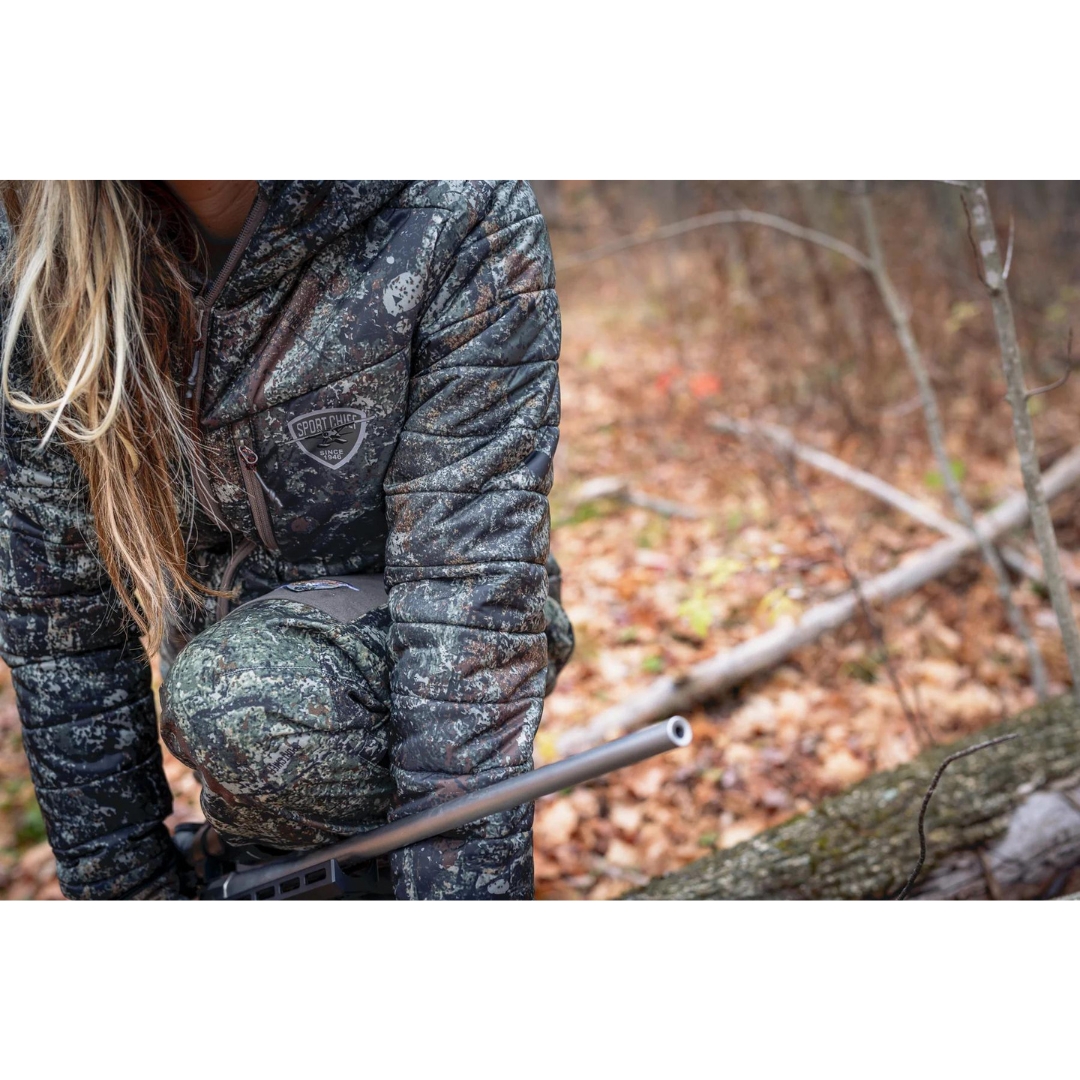 MANTEAU DE CHASSE SPORTCHIEF WILSON POUR FEMME COULEUR THE RIPPER VU DU MANTEAU À IMPRIMÉ CAMOUFLAGE TON DE TERRE PORTÉ PAR UNE FEMME ACCROUPIE DANS LA FORET