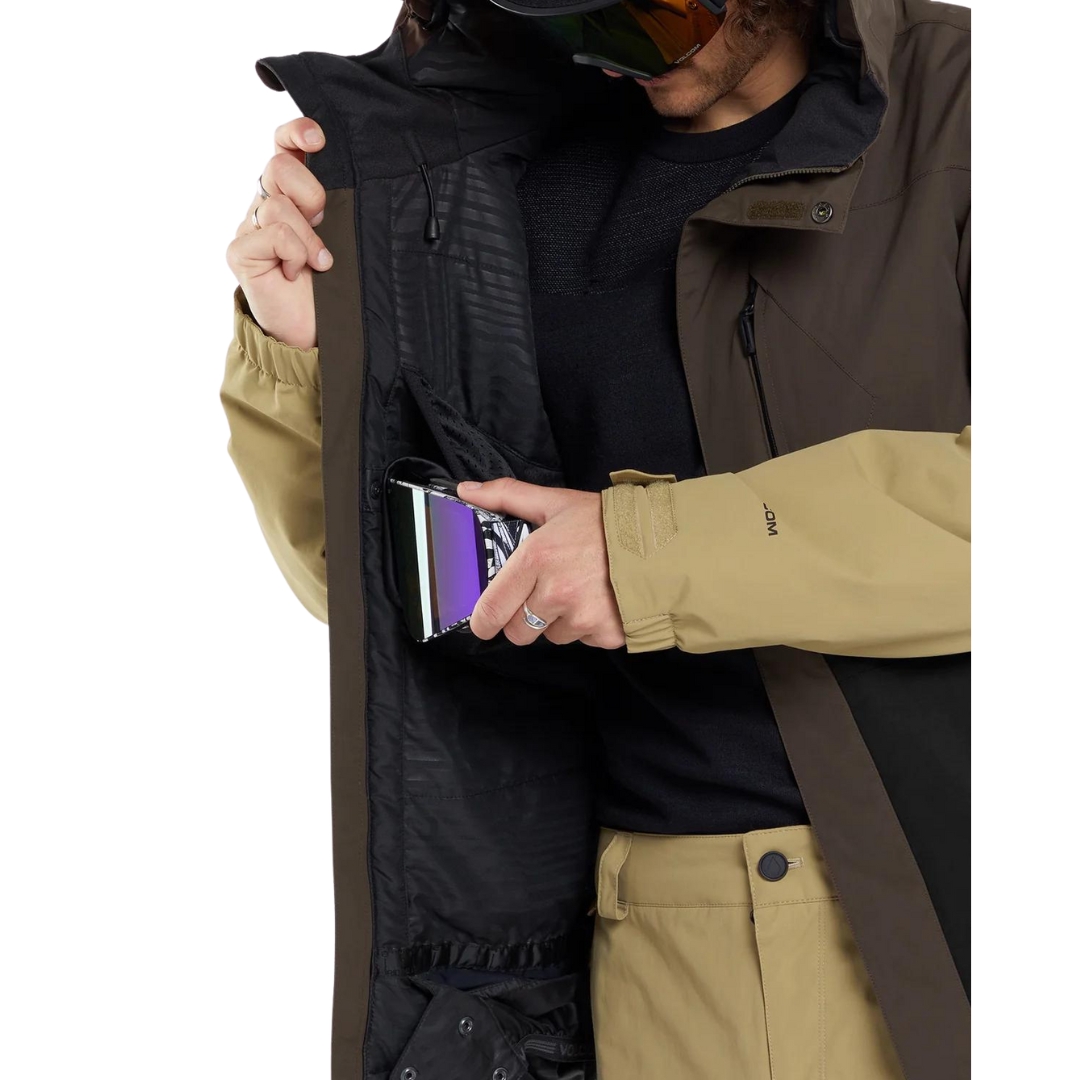 MANTEAU DE SKI EN GORETEX VOLCOM L POUR HOMME couleur brn-brown vu du manteau brun, noir et beige porté par un homme qui range ses lunettes de ski dans la poche prévu à cette fin