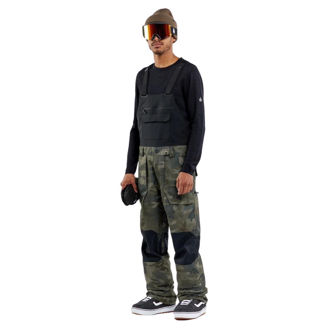 SALOPETTE DE SKI VOLCOM ROAN POUR HOMME couleur CWC-CLOUDWASH CAMO vue de la salopette à imprimé camouflage dans les ton de noir, brun et vert foret portée par un homme vu de la tête aux pieds de profil avant gauche