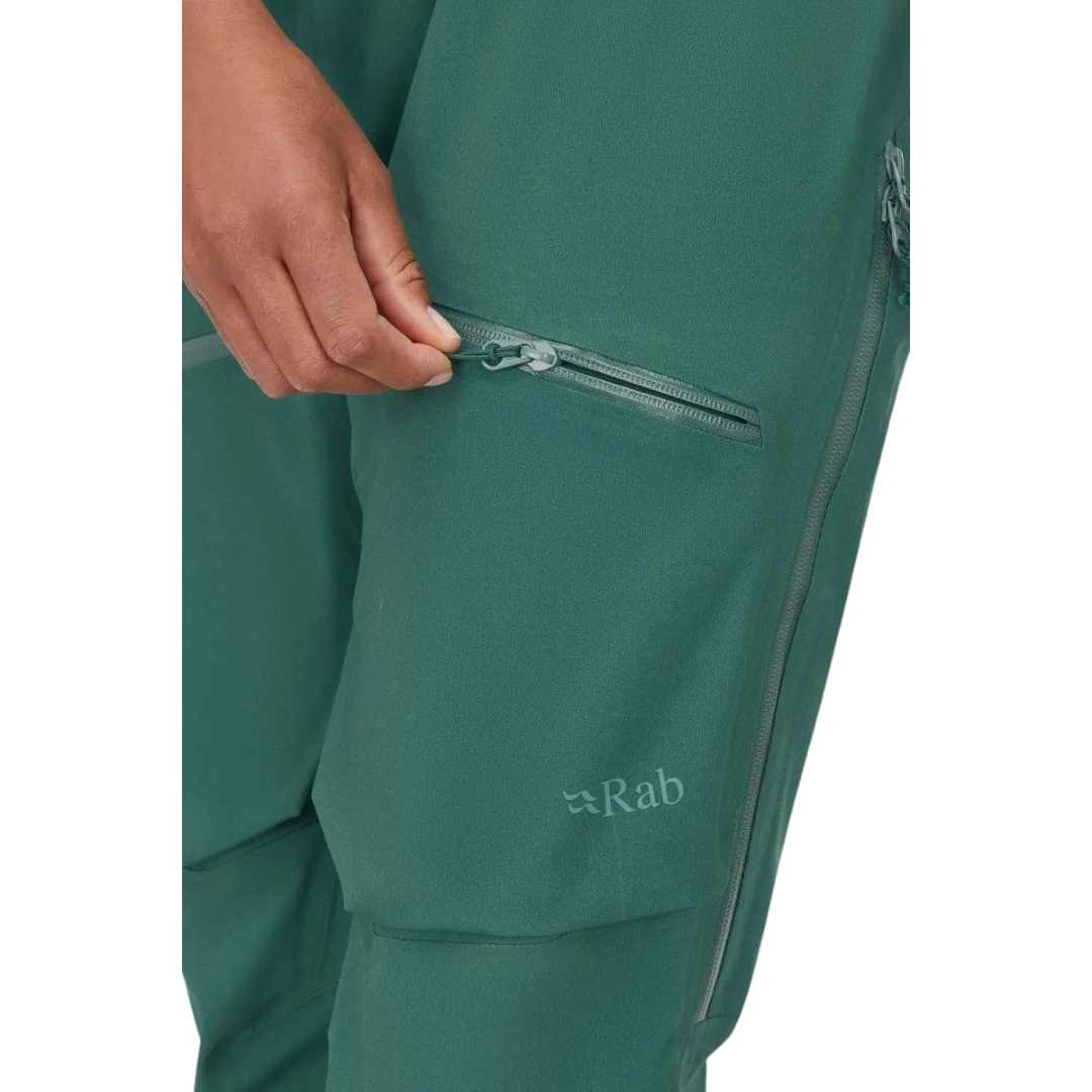 SALOPETTE IMPERMÉABLE DE SKI RAB KHROMA KINETIC POUR FEMME couleur green slate portée par une femme vue ouvrant la poche à fermeture éclair à la cuisse