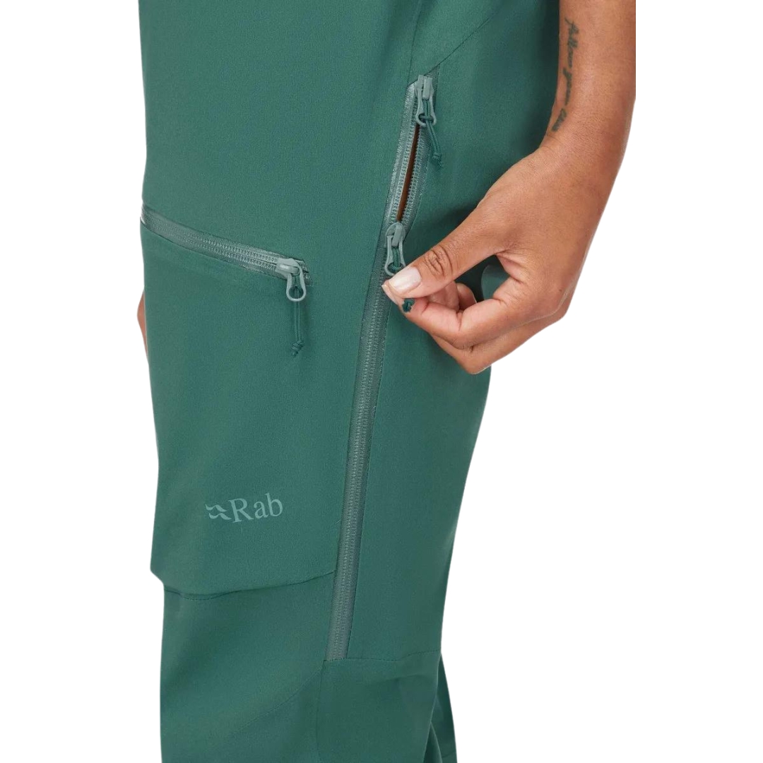 SALOPETTE IMPERMÉABLE DE SKI RAB KHROMA KINETIC POUR FEMME couleur green slate portée par une femme vue ouvrant la fermeture éclair sur le côté de la jambe