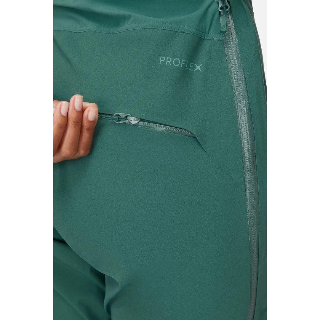 SALOPETTE IMPERMÉABLE DE SKI RAB KHROMA KINETIC POUR FEMME couleur green slate portée par une femme vue ouvrant la poche de fesse à fermeture éclair