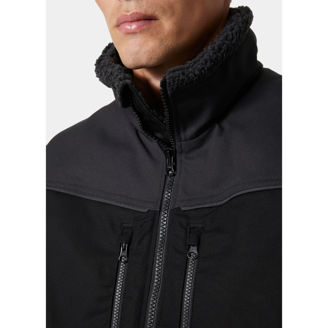 VESTE DE TRAVAIL DOUBLÉE SANS MANCHE HELLY HANSEN WORKWEAR OXFORD couleur black/ebony porté par un homme vu de face en gros plan sur le haut central de la veste entièrement noire on voit les deux textures des matières ainis que les fermeture éclaires aux poches de poitrine et celle de la fermture principale